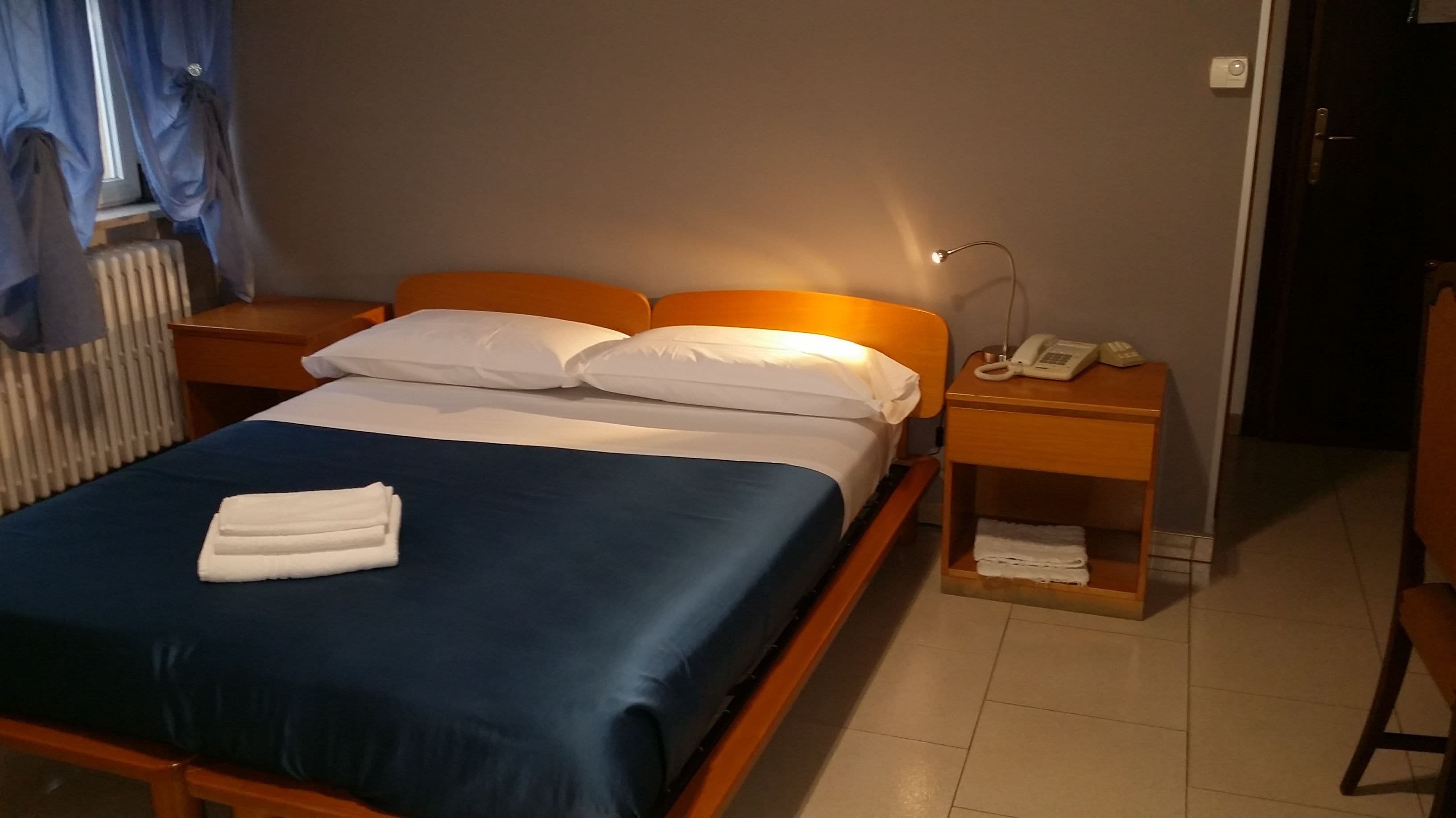 Una cama bien hecha con ropa de cama azul y almohadas blancas, una mesita de noche con lámpara y teléfono, y un radiador al fondo.