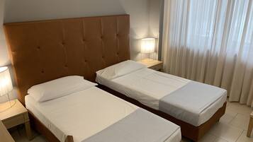 Quarto de hotel com duas camas de solteiro, cabeceira de madeira, luminárias laterais e janela com cortinas leves.