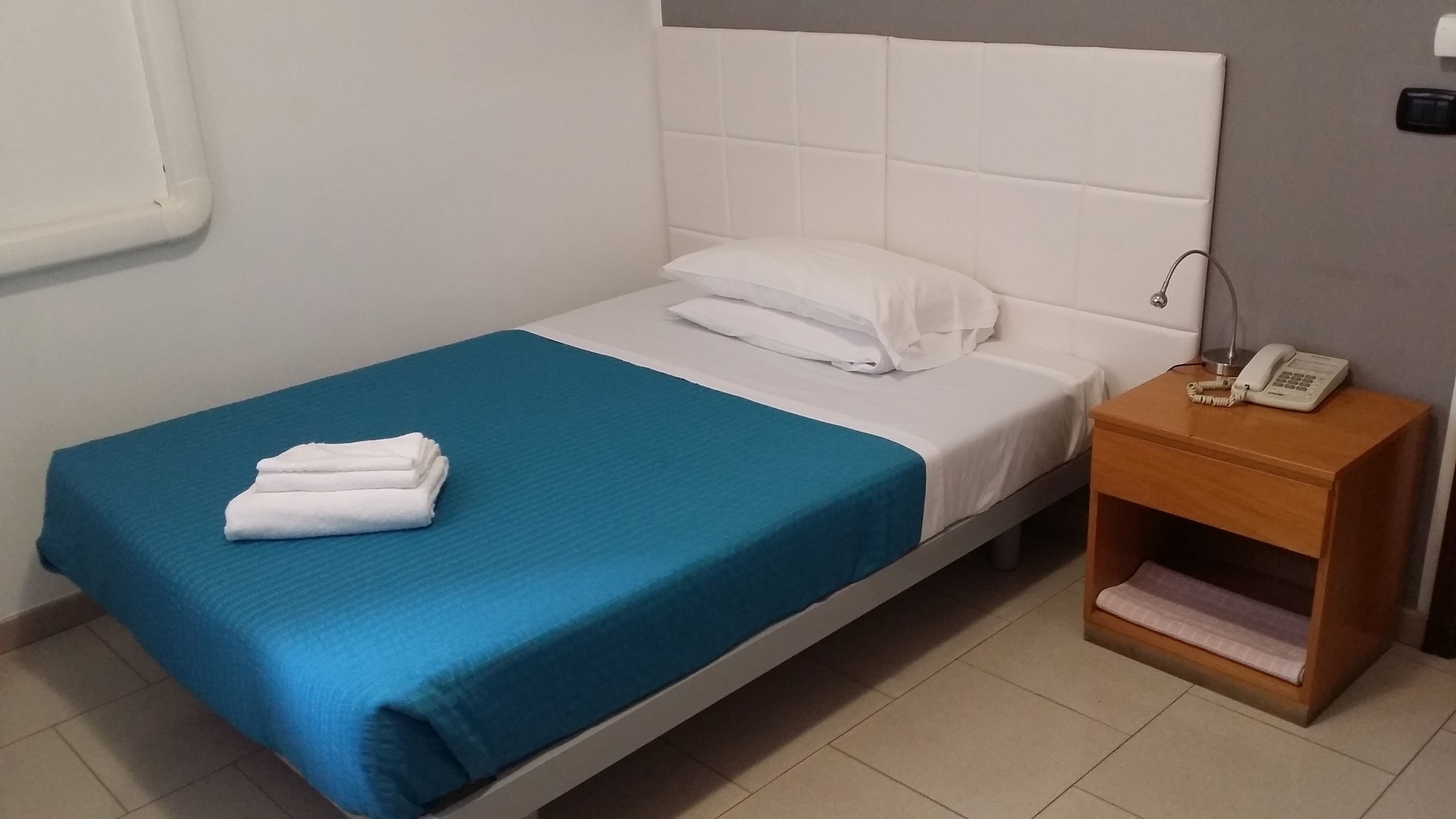 A single bed with a blue bedspread and white pillows, a wooden nightstand with a telephone, and a small shelf with a pink towel.