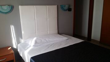 A neatly made bed with white linens, a dark patterned bedspread, and a wooden nightstand with a drawer.