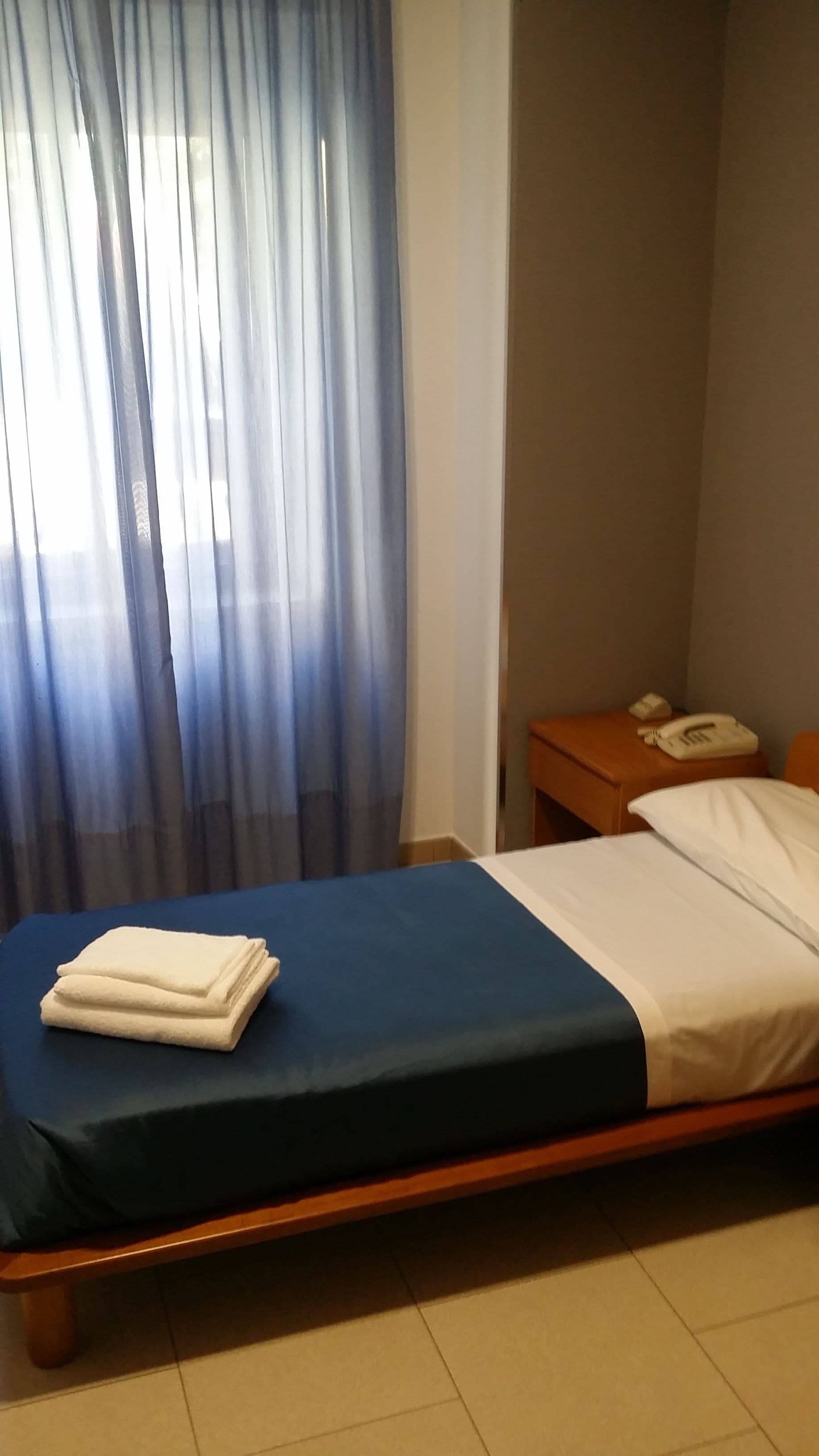 Une chambre d’hôtel avec un lit, une table de chevet avec un téléphone et une fenêtre avec des rideaux légers.