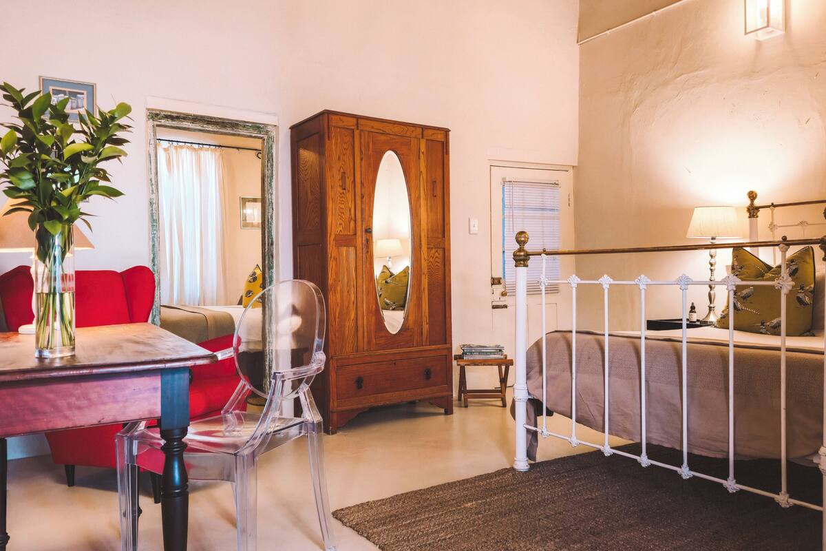 A bedroom with a wooden wardrobe, a bed with white metal headboard, a red chair, a clear glass table, and a vase with green plants.