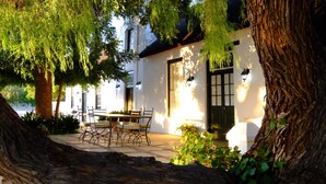 A white building with a black door and windows, surrounded by trees and a patio with a table and chairs. - Onse Rus Guest House (Prince Albert)