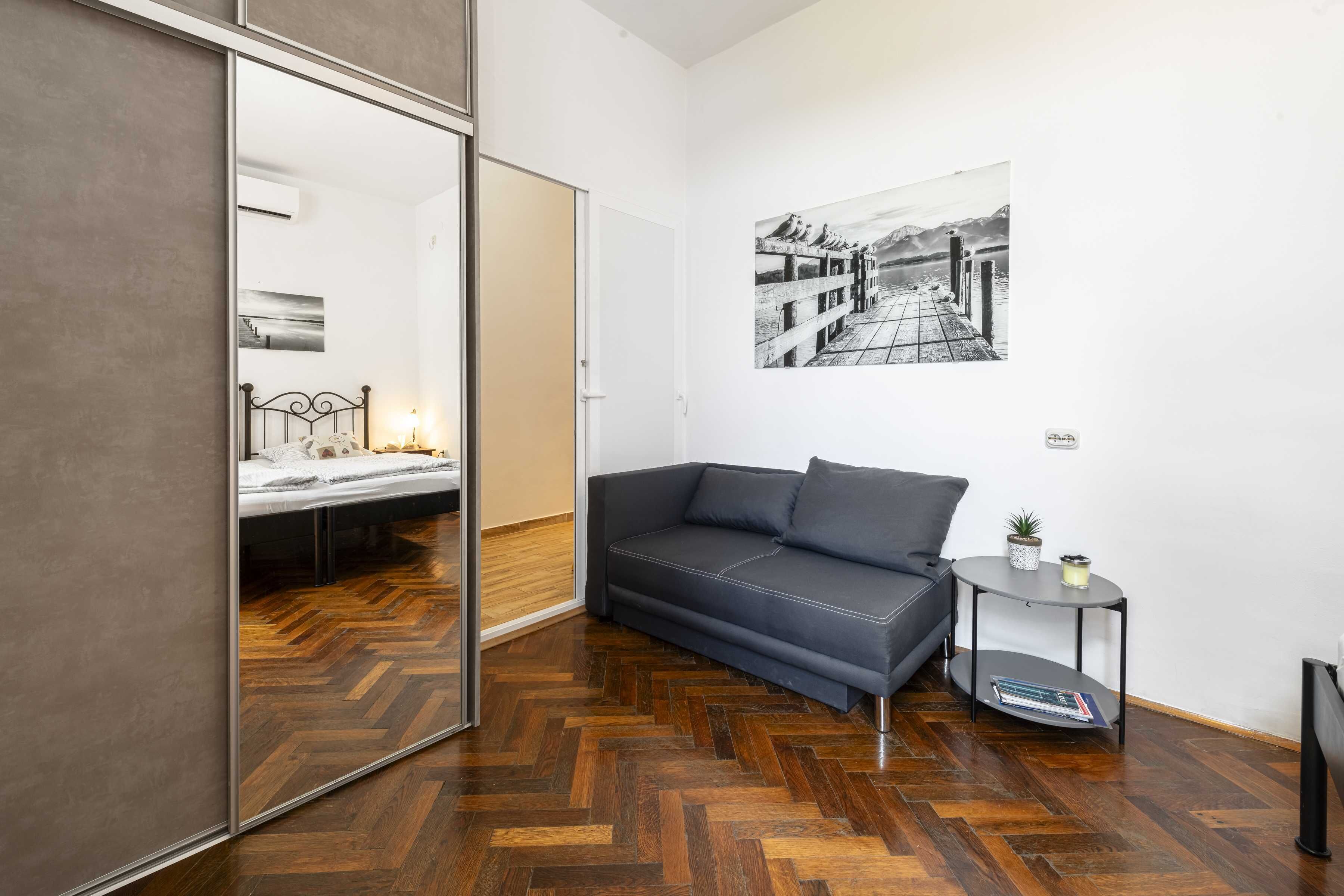 A modern room with a herringbone wooden floor, a grey sofa, a small round table with a plant, and a large mirror reflecting a bed with a nightstand.