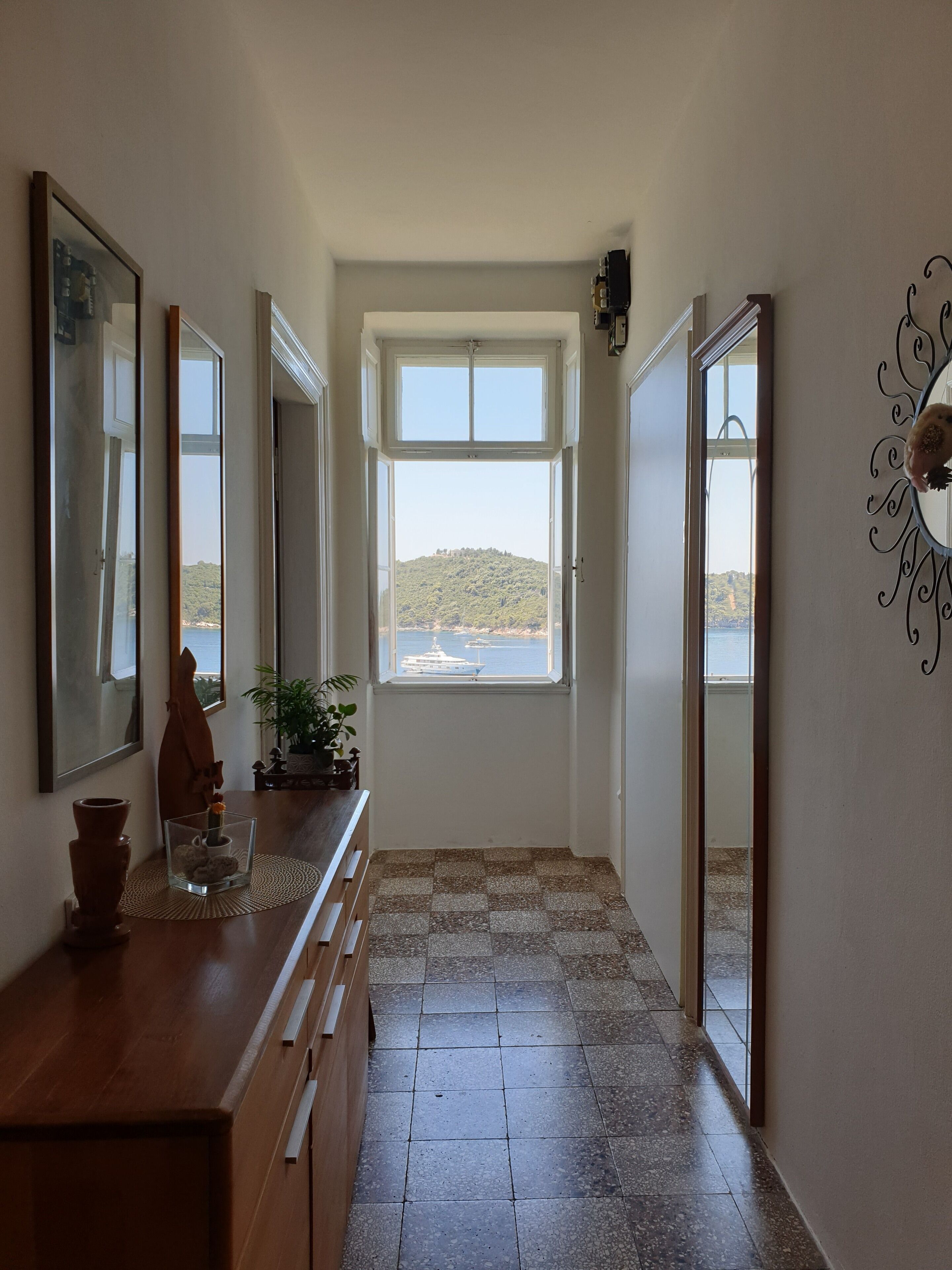 A hallway with a view of a body of water and a hill. There is a wooden bench, a vase with flowers, and a potted plant.