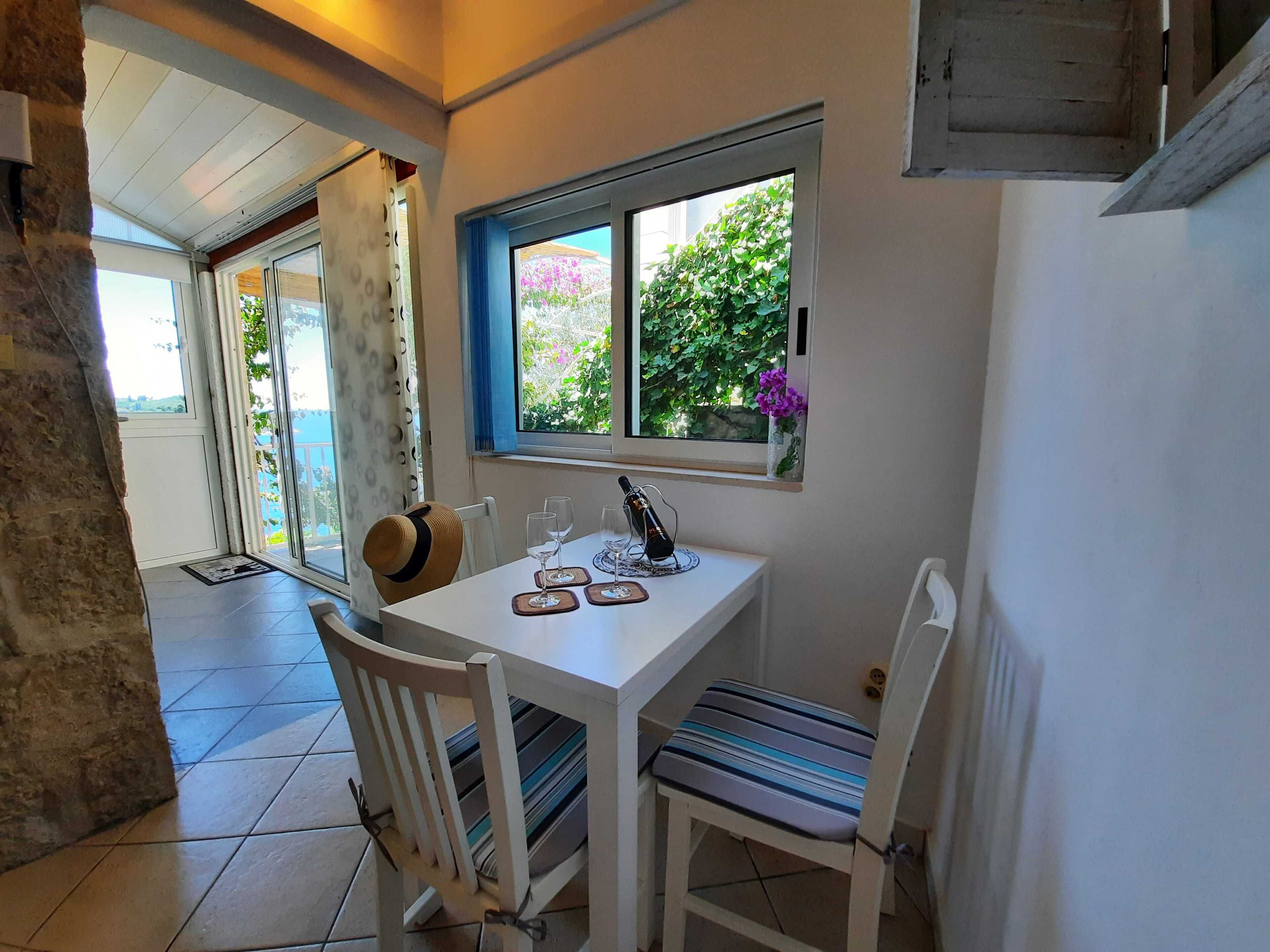 Uma área de refeições com mesa branca, duas cadeiras, janela com vista para vegetação e um vaso com flores.