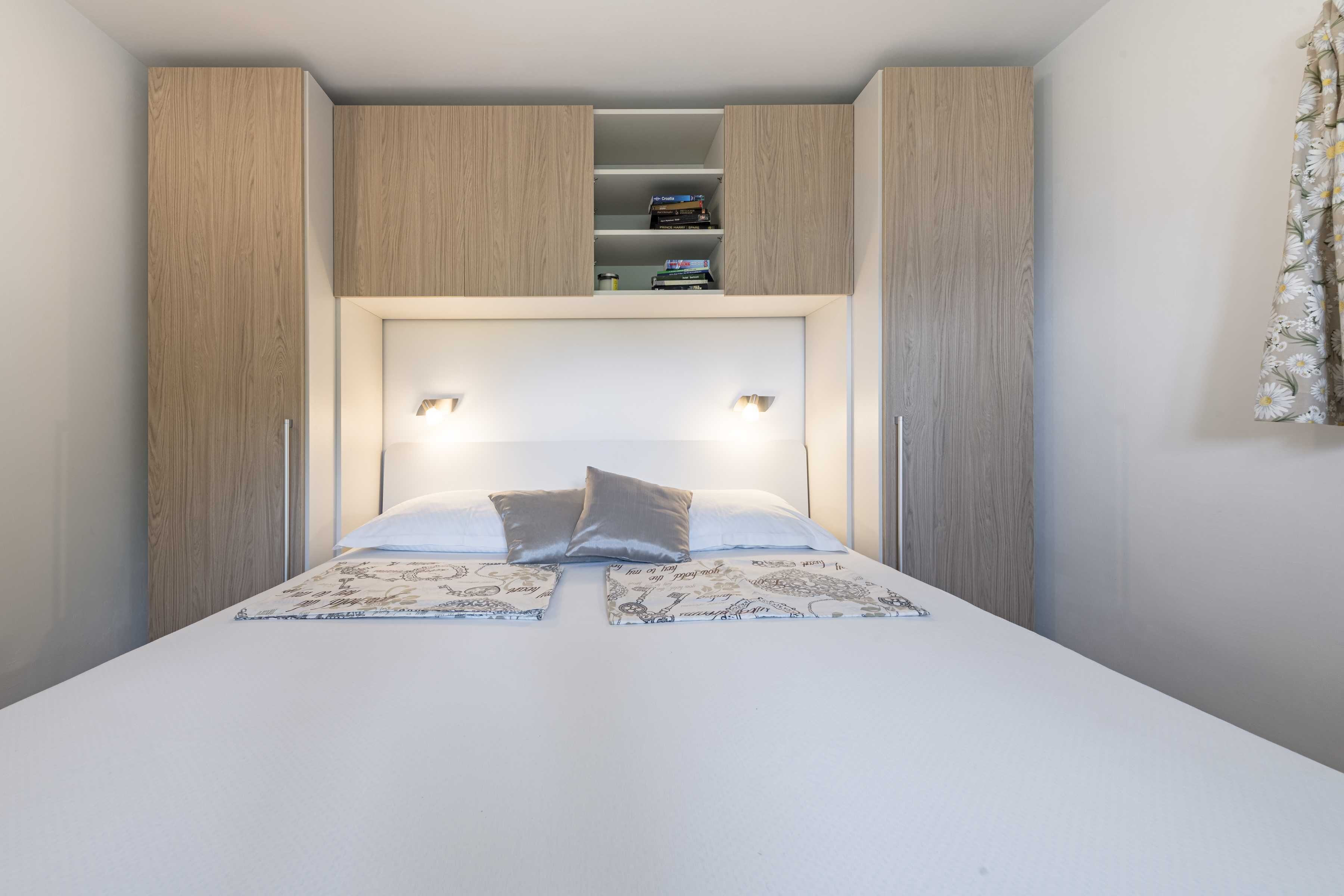 Un dormitorio con cama, dos mesitas de noche, un armario y una estantería sobre la cama.