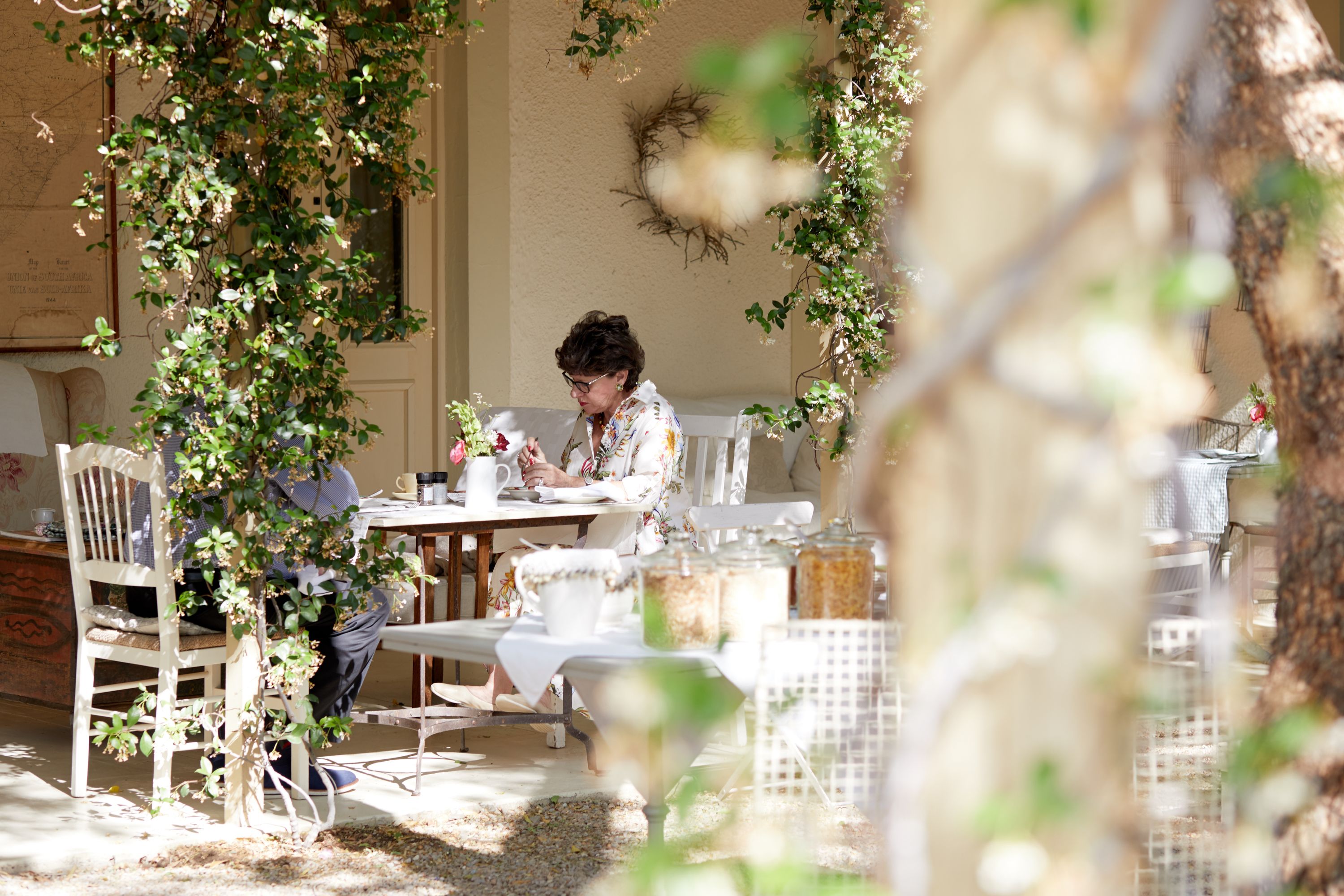 Un espace extérieur aménagé pour les repas, avec des chaises et des tables blanches, entouré de verdure et agrémenté d’un mur décoratif.