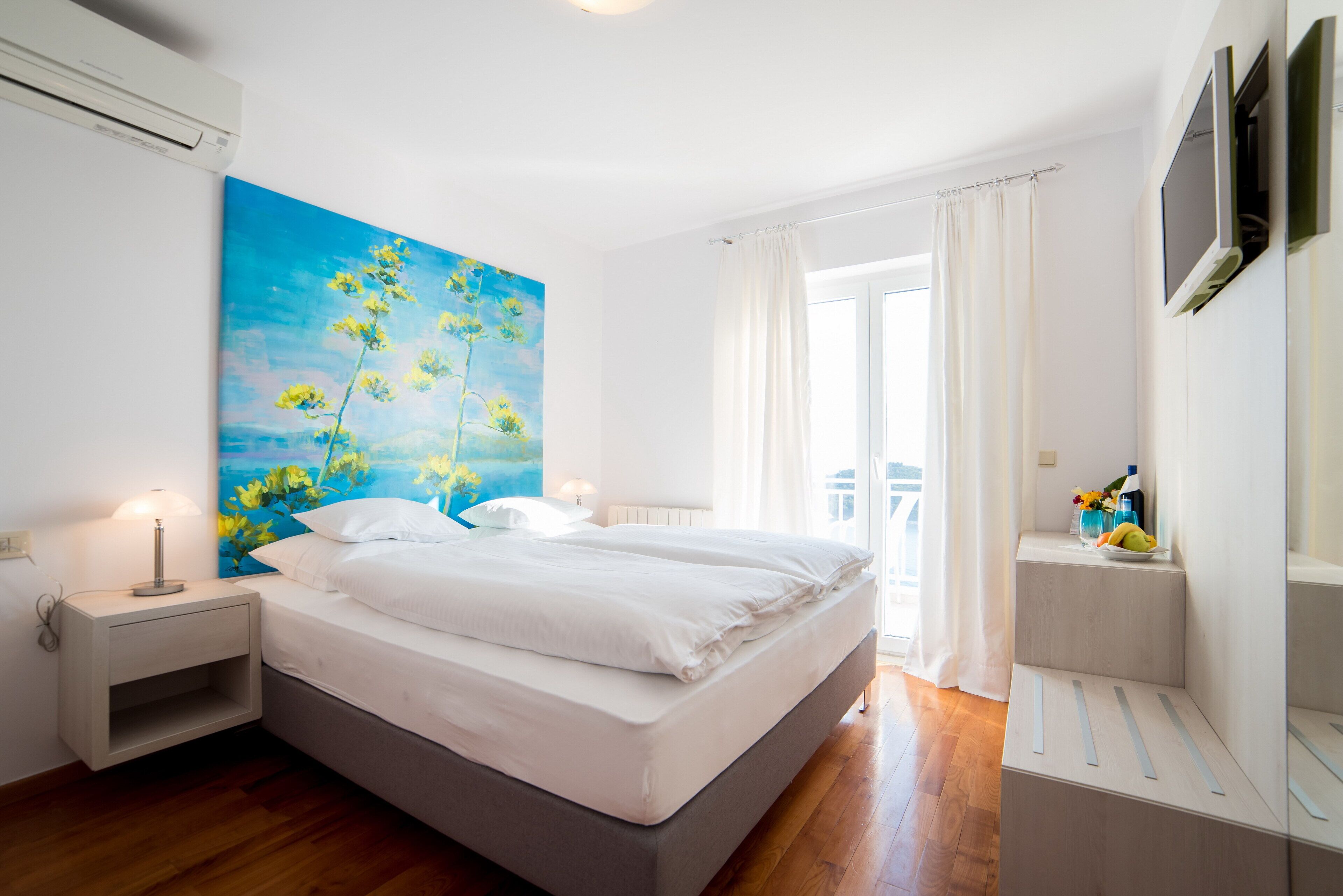Un dormitorio con cama, mesita de noche, un cuadro, televisor y ventana con cortinas.