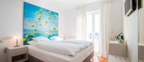 Un dormitorio con cama, mesita de noche, un cuadro, televisor y ventana con cortinas.