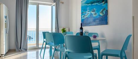 A dining area with teal chairs and a table, a painting of ocean waves on the wall, and a view of the sea through the glass door.