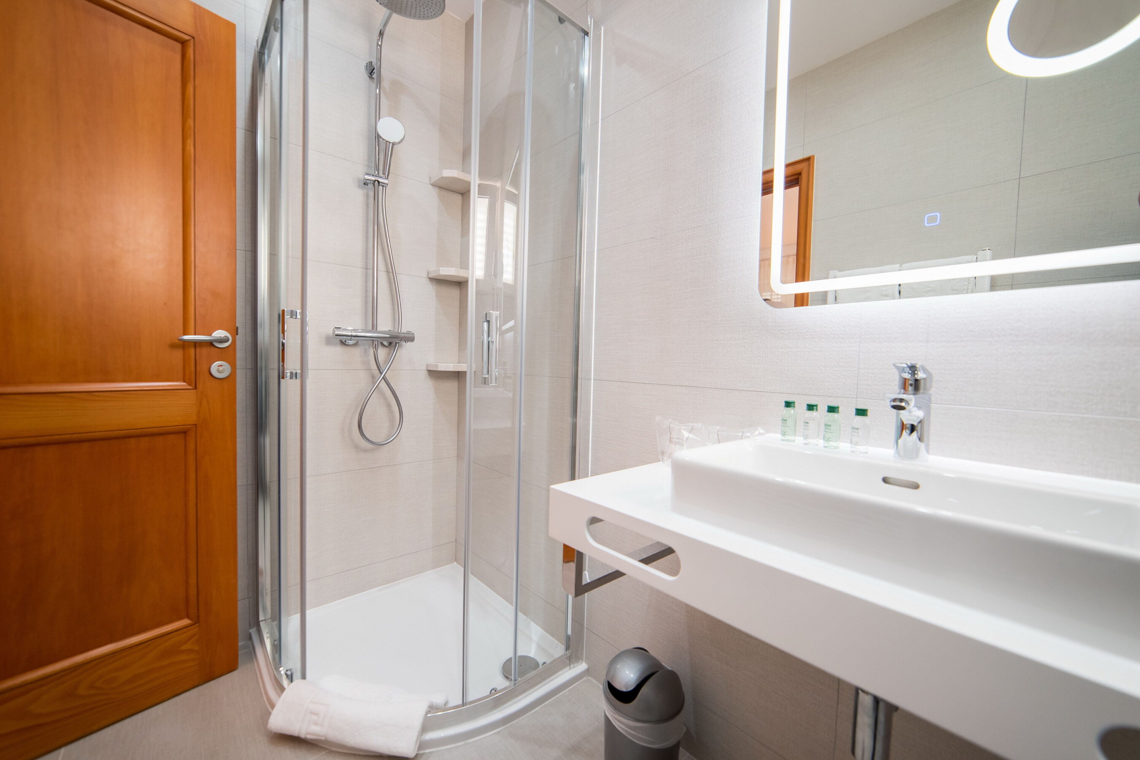 Un baño moderno con cabina de ducha de vidrio, puerta de madera, lavabo blanco con espejo y artículos de aseo sobre el lavabo.