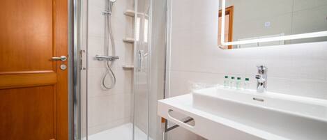 Un baño moderno con cabina de ducha de vidrio, puerta de madera, lavabo blanco con espejo y artículos de aseo sobre el lavabo.