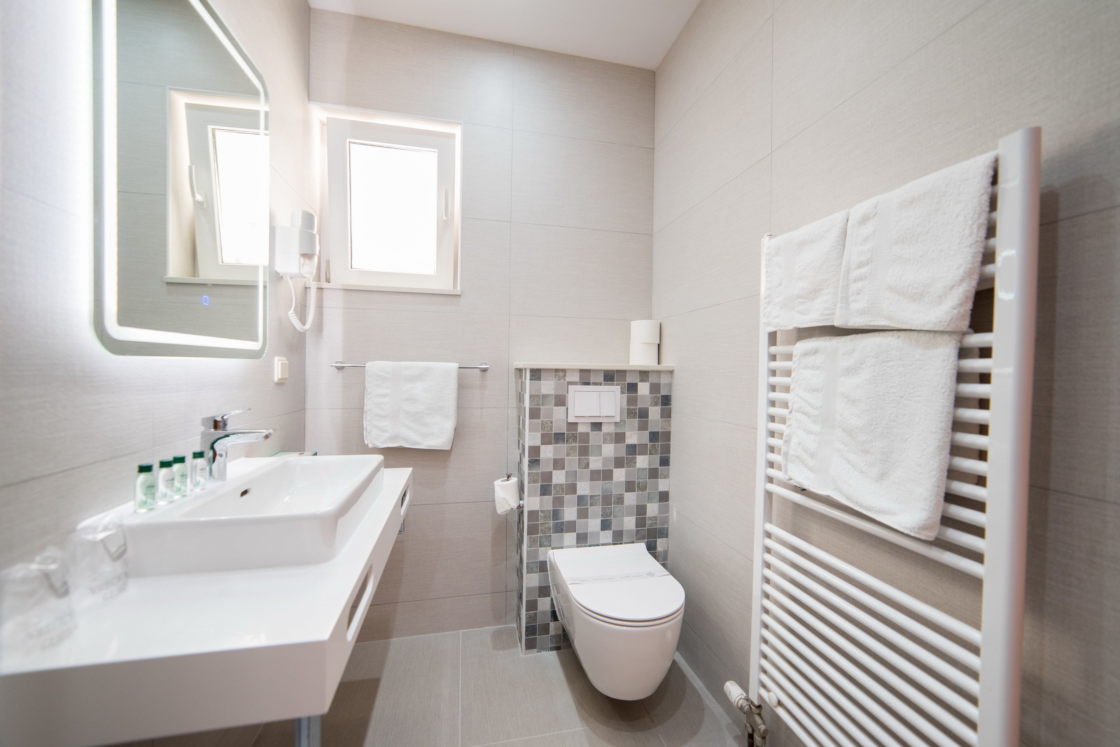 A modern bathroom with a white sink, mirror, and toilet. There are towels hanging on a rack and a tiled shower wall.