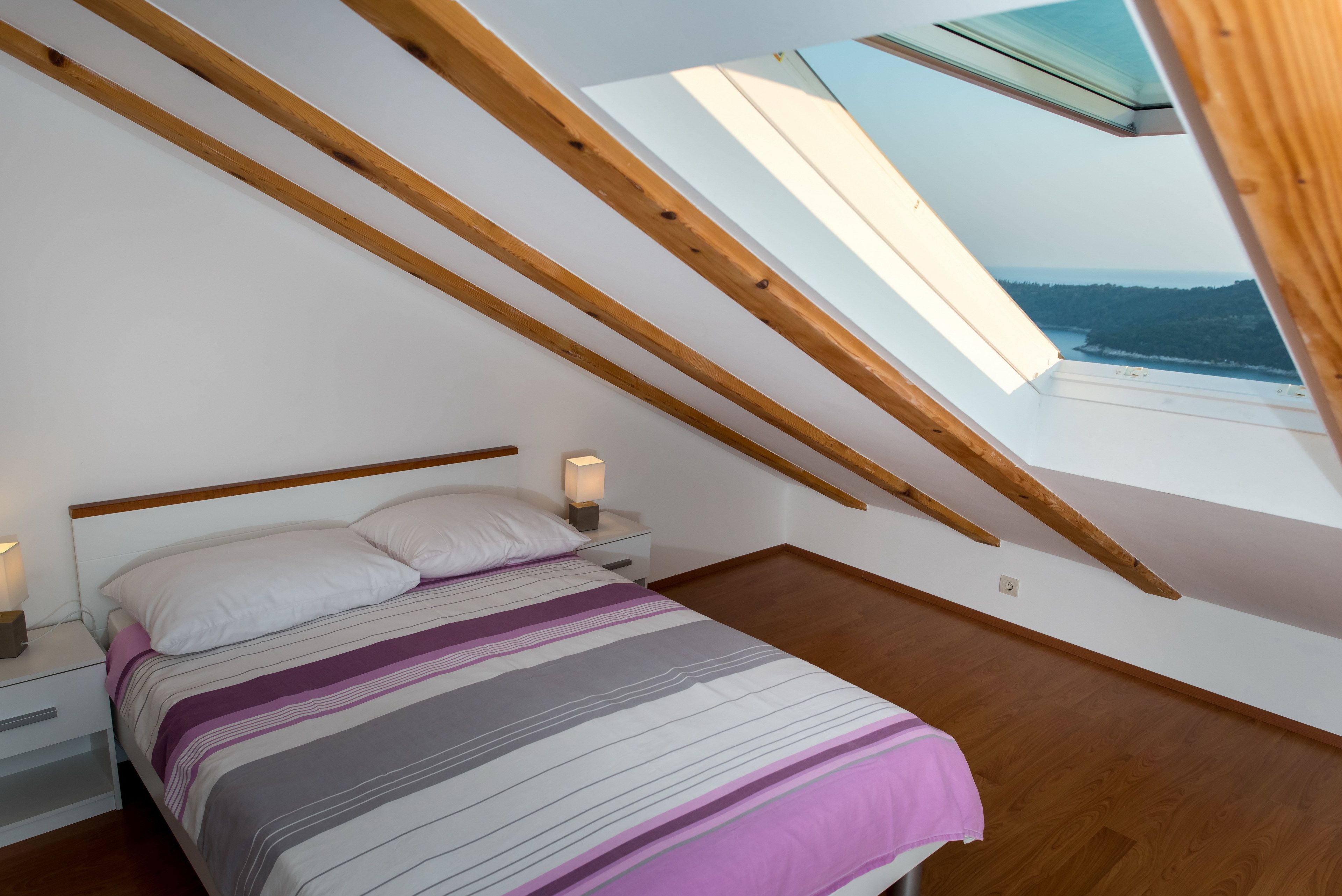 A bedroom with a sloped ceiling, a bed with a striped blanket, bedside lamps, and a view through a skylight.