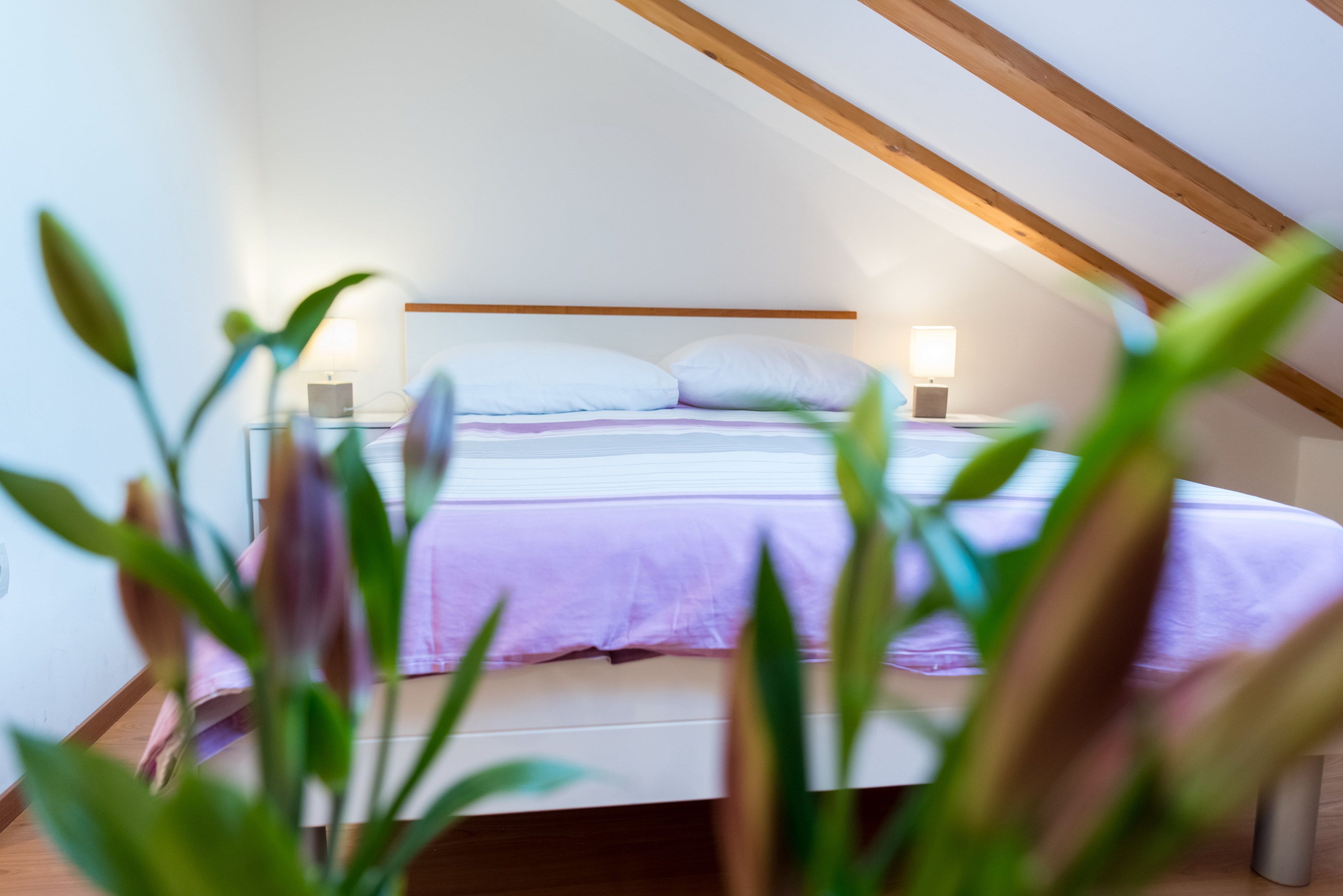 Un dormitorio con techo inclinado, una cama con ropa de cama blanca y morada, dos lámparas de noche y un cabecero de madera.