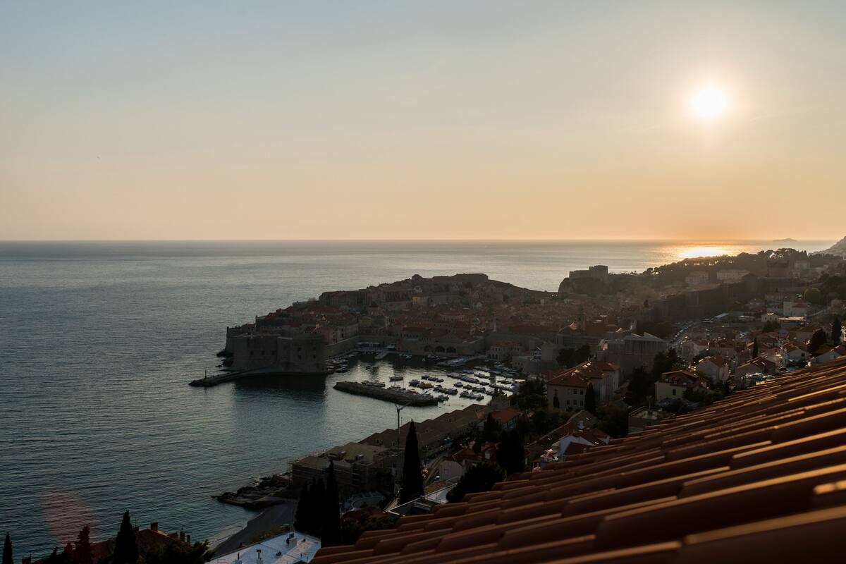 A coastal cityscape with a prominent historic building by the sea, a marina, and a hilly backdrop.