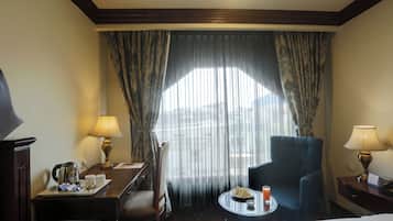 A hotel room with a bed, a desk with a kettle, a chair, a small round table with a drink and a plate of sandwiches, and a window with sheer curtains.