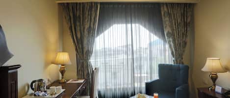 A hotel room with a bed, a desk with a kettle, a chair, a small round table with a drink and a plate of sandwiches, and a window with sheer curtains.