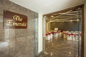 A hotel entrance with a sign reading 'The Emerald,' a glass door, and rows of white chairs with red sashes. - Vesta Sapphire Jaipur - A City Center Hotel (Jaipur)