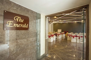Entrada de un hotel con un cartel que dice 'The Emerald', puerta de cristal y filas de sillas blancas con cintas rojas.