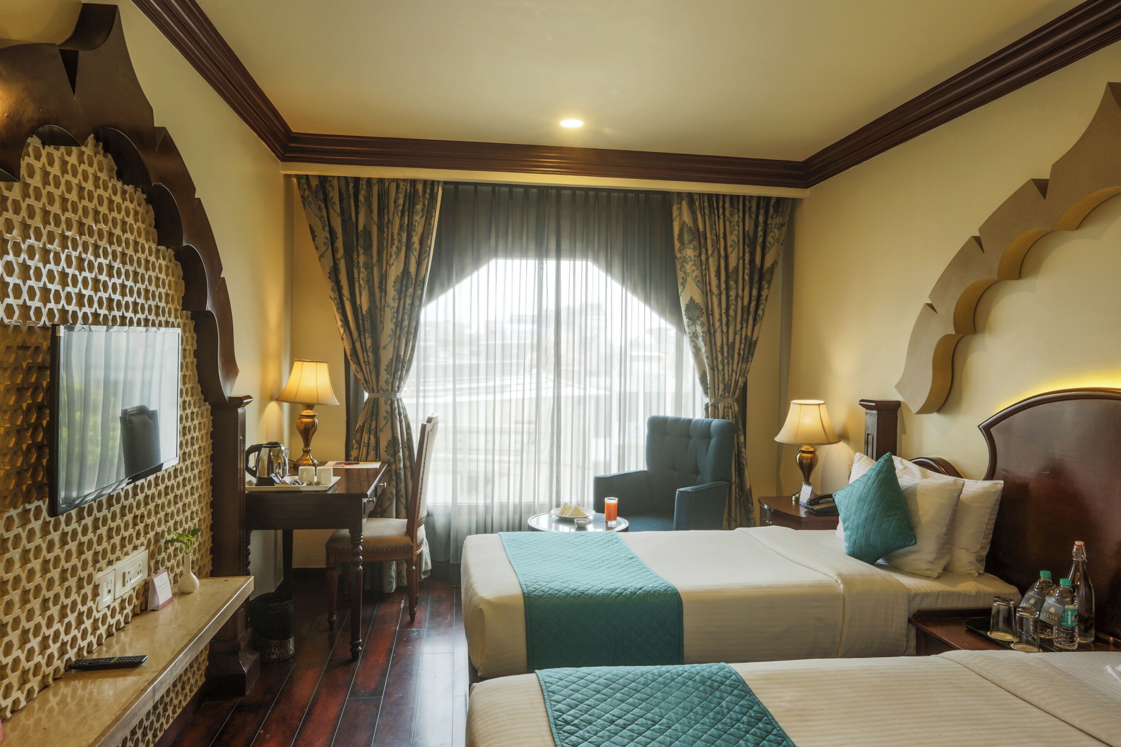 A hotel room with two beds, a desk, a chair, a table with a lamp, and a large window with curtains.