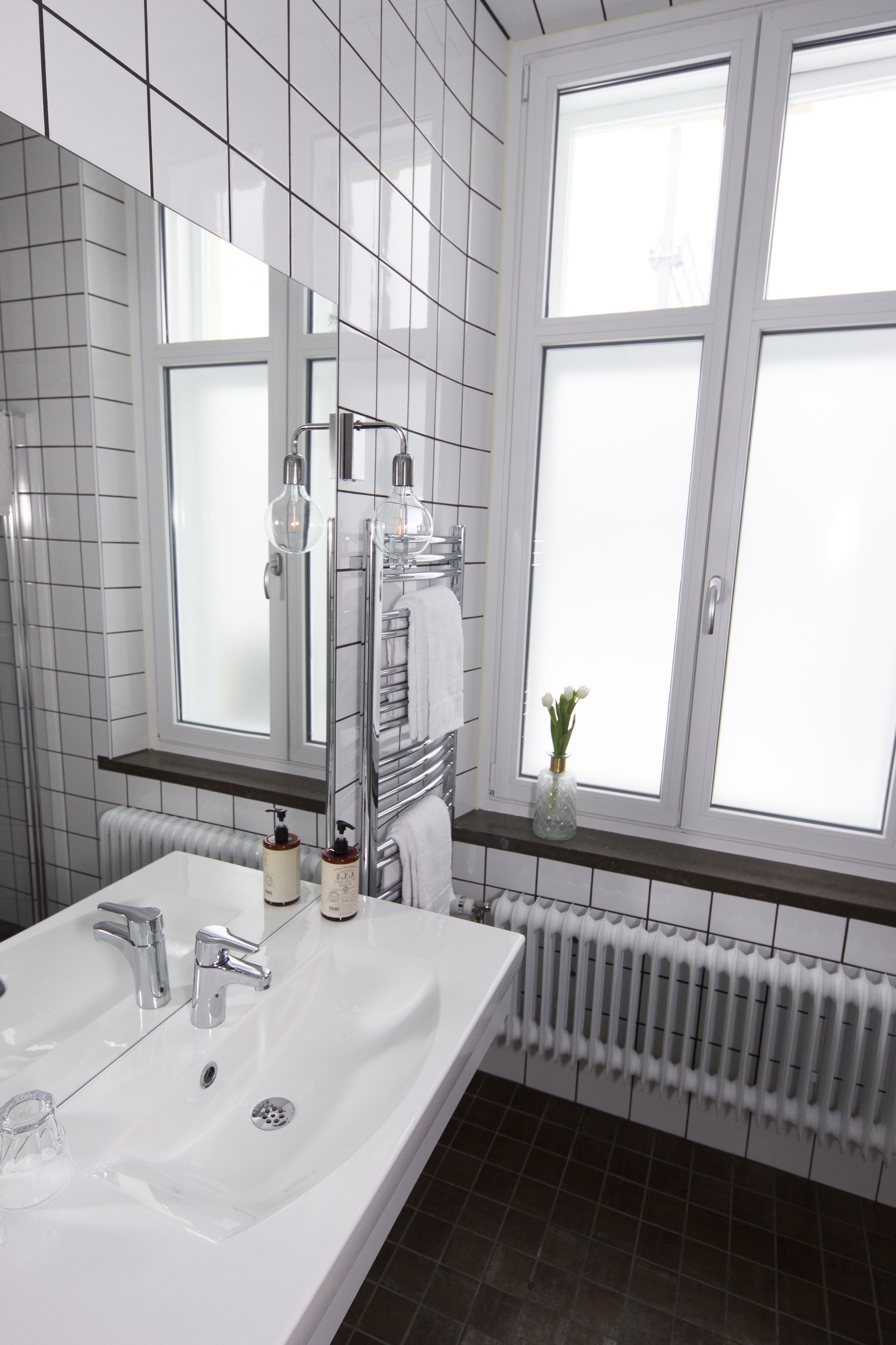 Un baño con lavamanos blanco, dos llaves, un espejo, toallero, ventana y radiador.