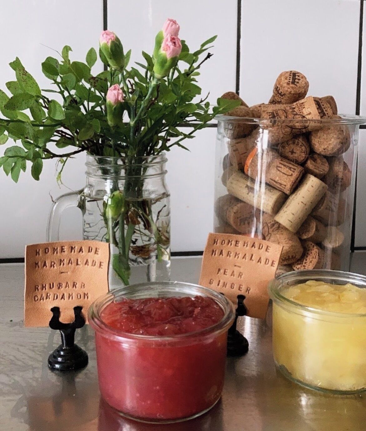 Una mesa con un tarro de vidrio con líquido rosa, un tarro de vidrio con líquido amarillo, un florero de vidrio con flores rosas y un tarro de tapones de corcho.