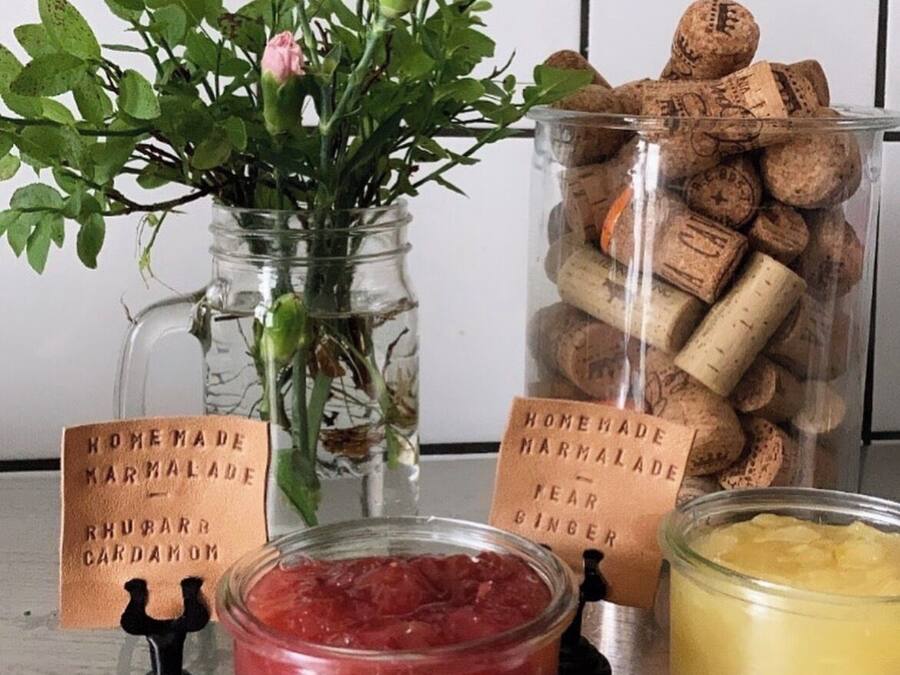 Una mesa con un tarro de vidrio con líquido rosa, un tarro de vidrio con líquido amarillo, un florero de vidrio con flores rosas y un tarro de tapones de corcho.