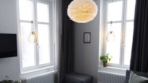 A modern living room with a grey sofa, two black stools, a hanging light fixture, and large windows with curtains. - Livin Station (Örebro)