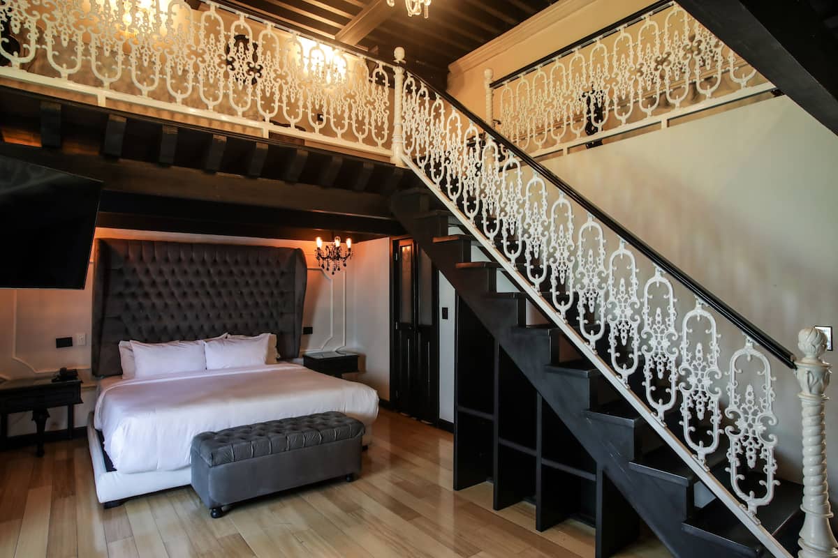 A bedroom with a large bed, a nightstand, a bench, and a staircase with ornate railings.