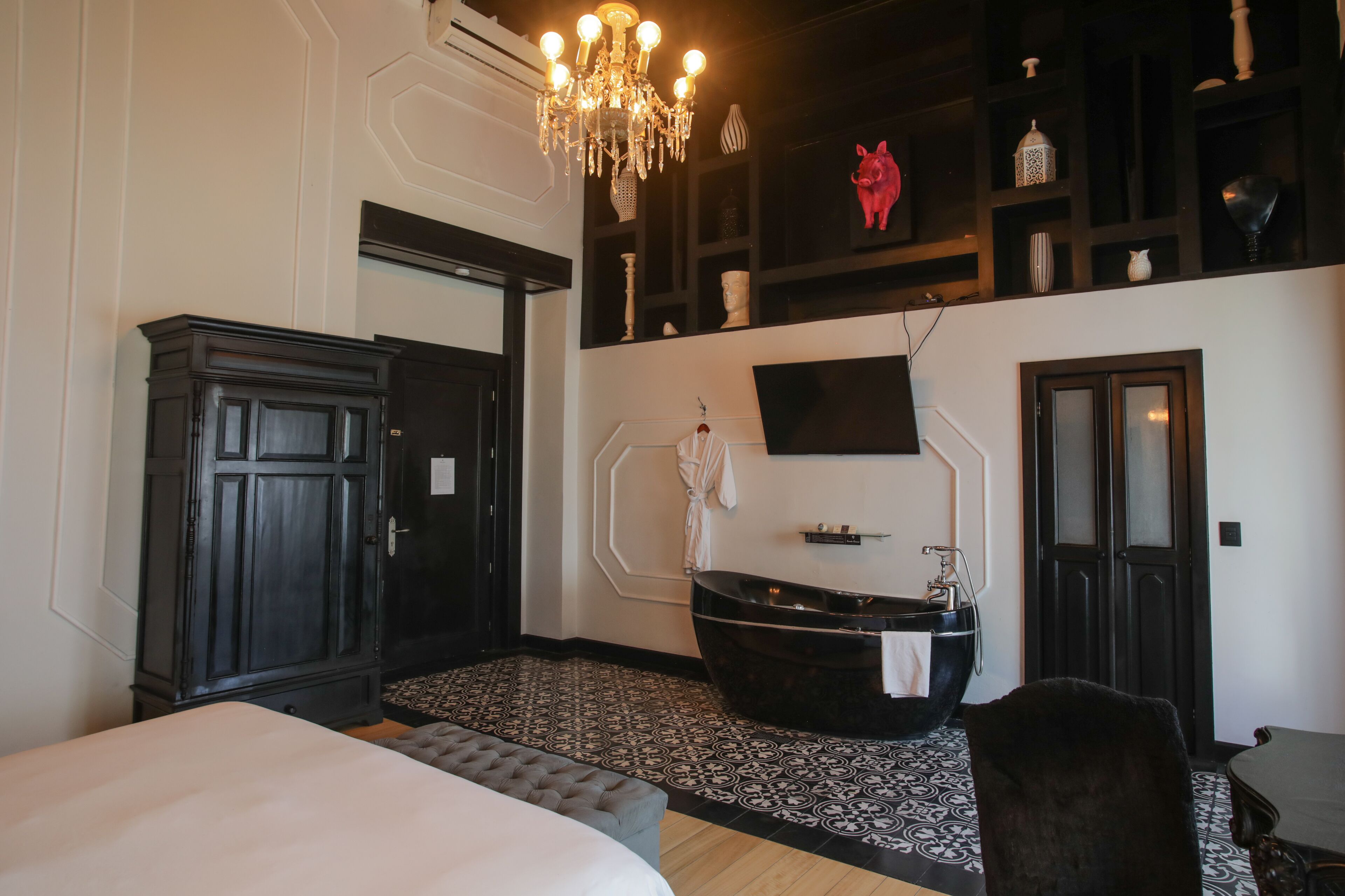 A bedroom with a large bed, a black bathtub, a chandelier, and a TV mounted on the wall.