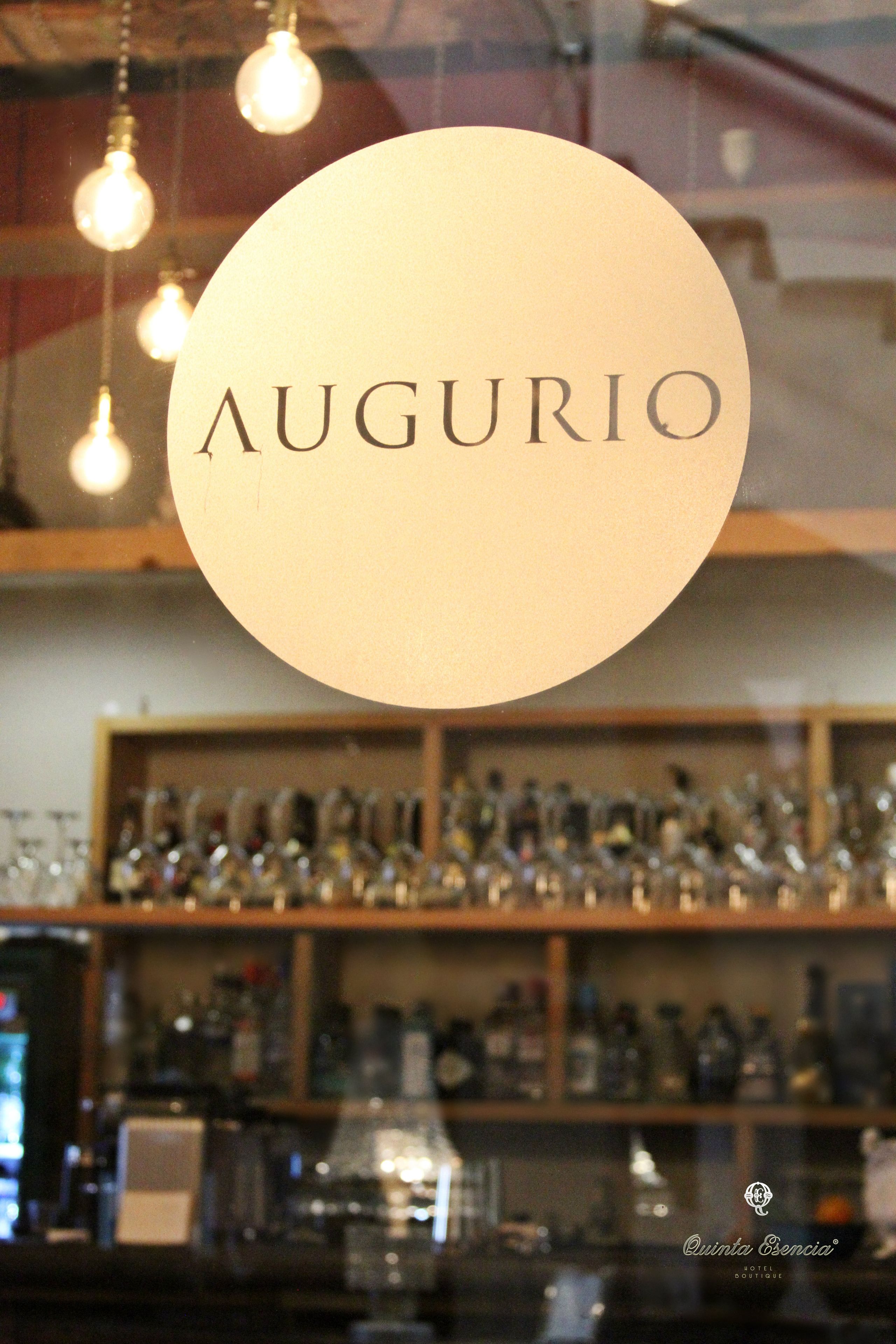Un letrero circular con la palabra "AUGURIO" en el centro, colgando sobre una zona de bar con estantes llenos de botellas y vasos.
