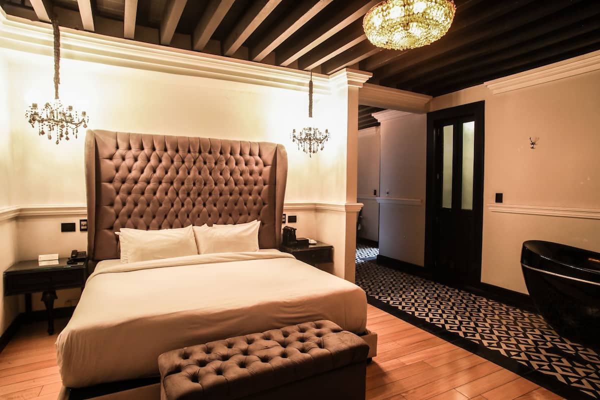 A hotel room with a large bed, a tufted headboard, two bedside tables, a chandelier, and a patterned floor.