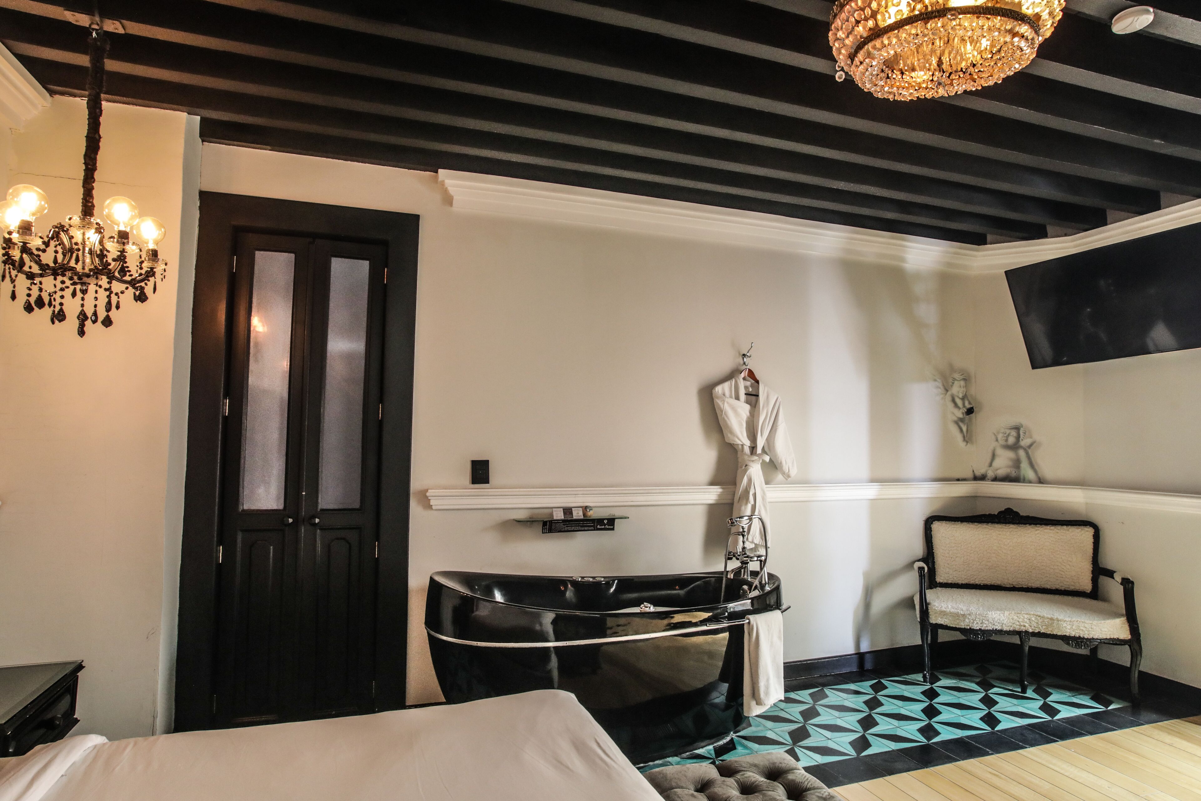 Habitación de hotel con una bañera negra, una banca, un candelabro y un televisor montado en la pared.
