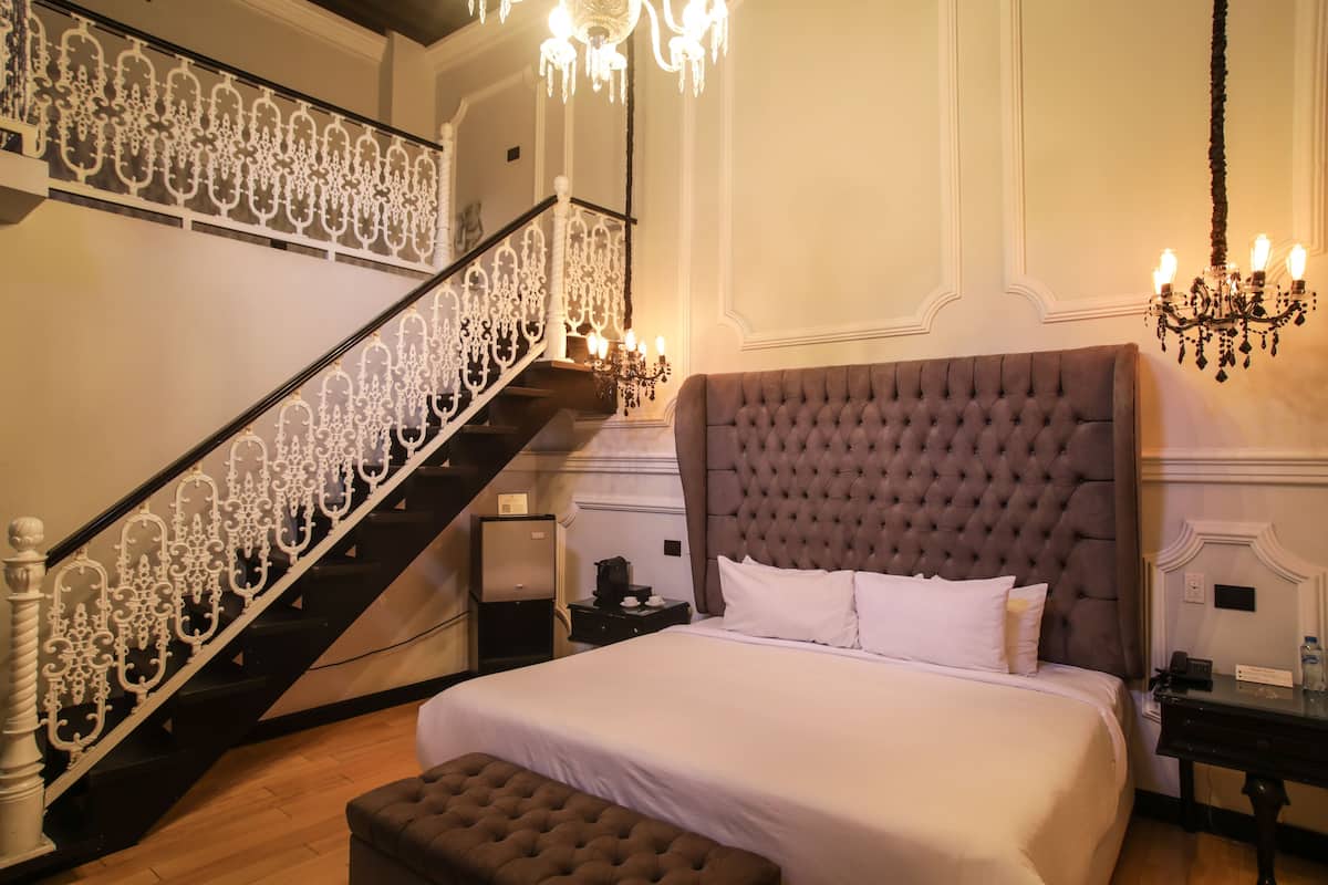 A bedroom with a large bed, a chest of drawers, a nightstand, a dresser, a chandelier, and a staircase with ornate railings.