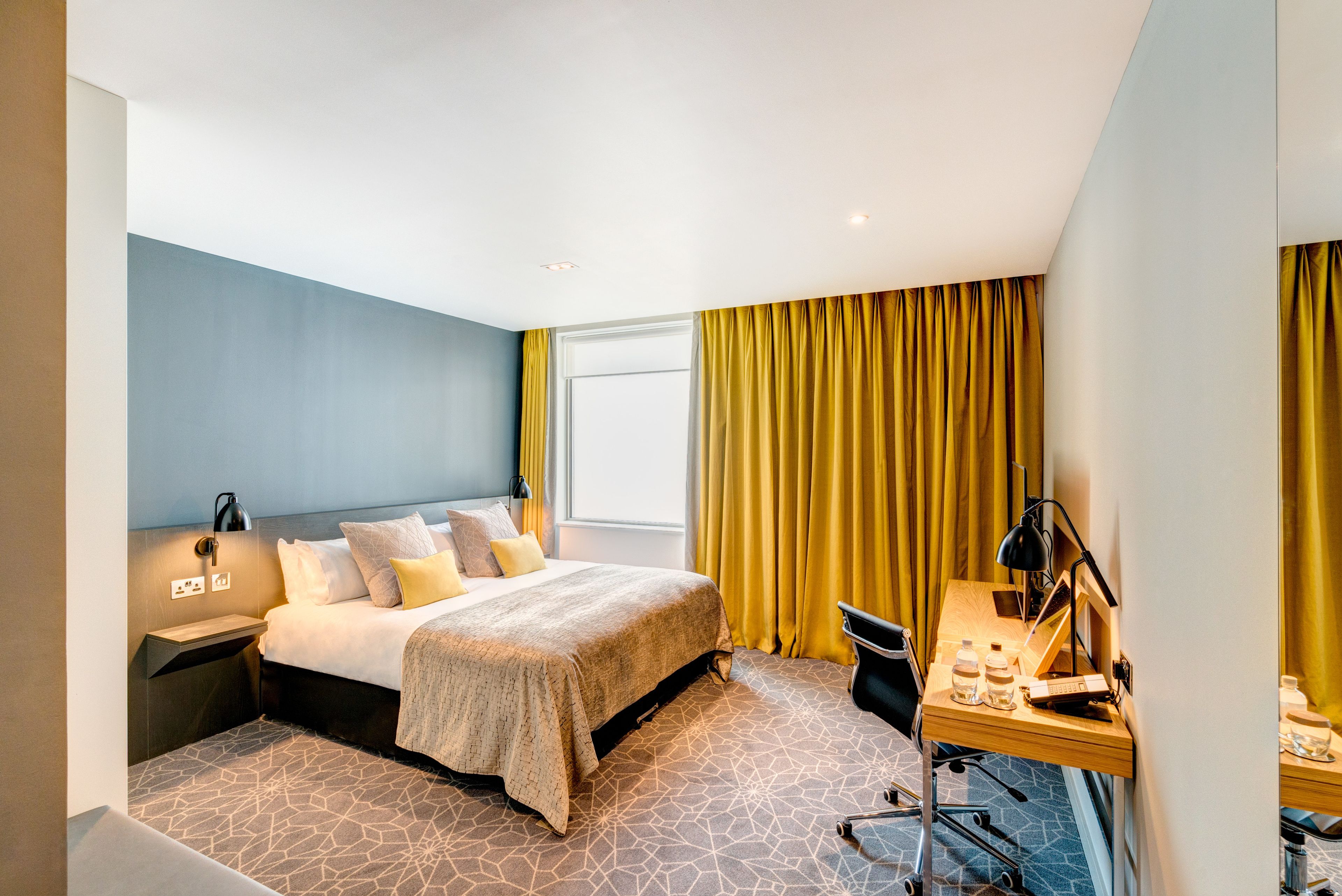 A modern hotel room with a bed, desk, and a view of the outdoors.