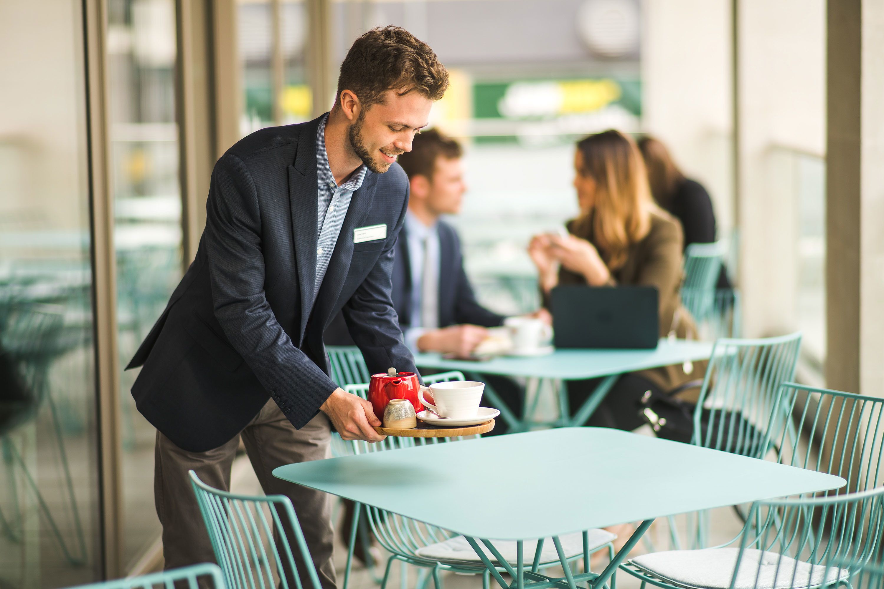 A waiter serving coffee to a table in a modern cafe with glass walls and green chairs.