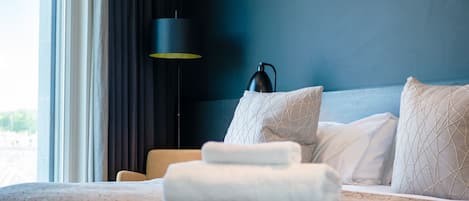 Een net opgemaakt bed met een opgerolde handdoek, een zwarte vloerlamp en uitzicht op de omgeving door een raam.