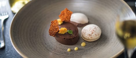Un plato de postres con diferentes elementos, incluyendo una esfera de chocolate, una esfera blanca y un macaron, acompañado de una copa de vino.