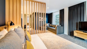 A modern hotel room with a large bed, a seating area with a sofa and a TV, and a wooden partition. - Apex City of Bath Hotel (Bath)
