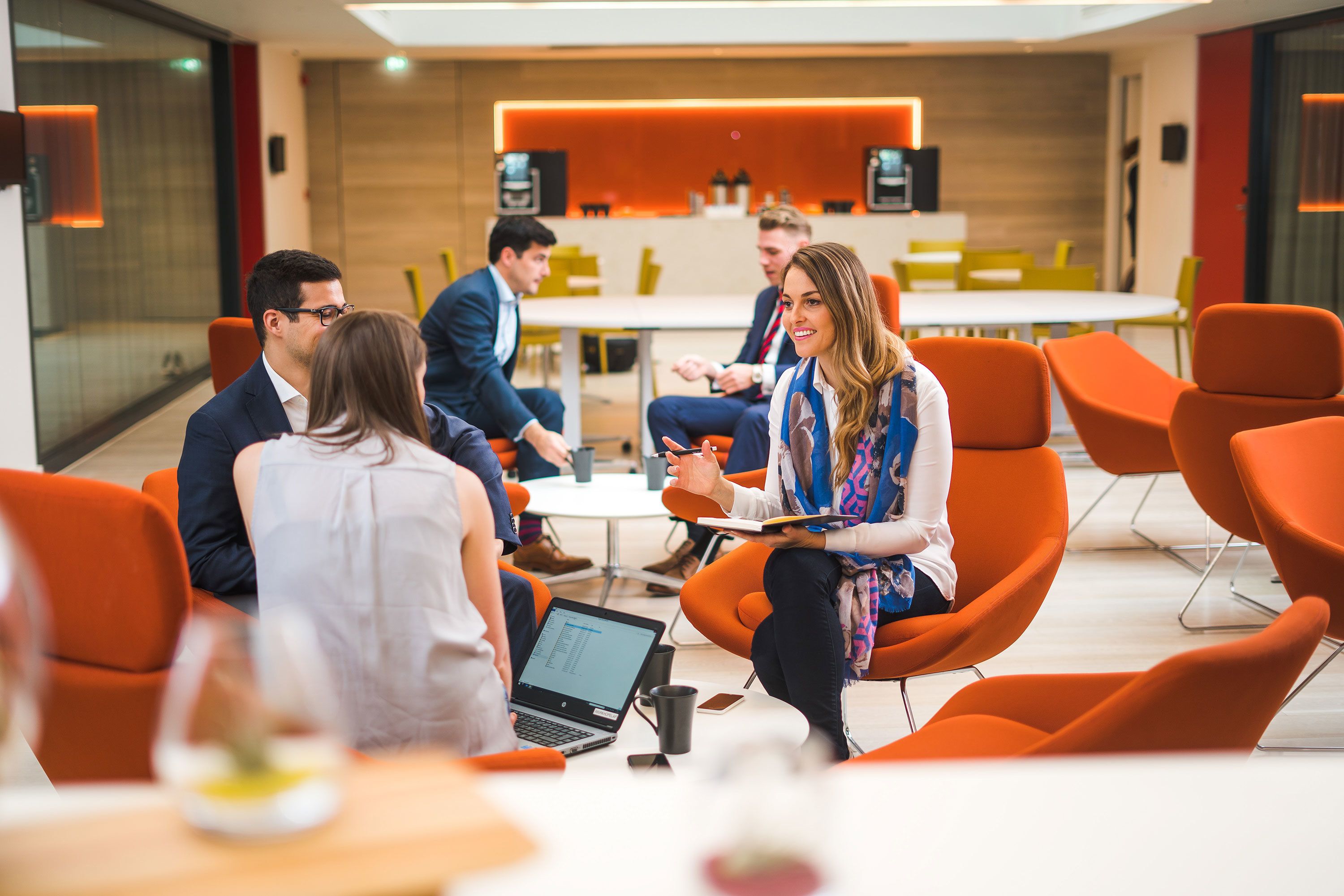 A modern meeting room with orange chairs, a round table, and a laptop on the table.