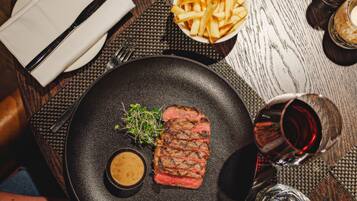 A steak with a side of fries and a small bowl of sauce on a black plate, accompanied by a glass of red wine and a napkin on a wooden table.
