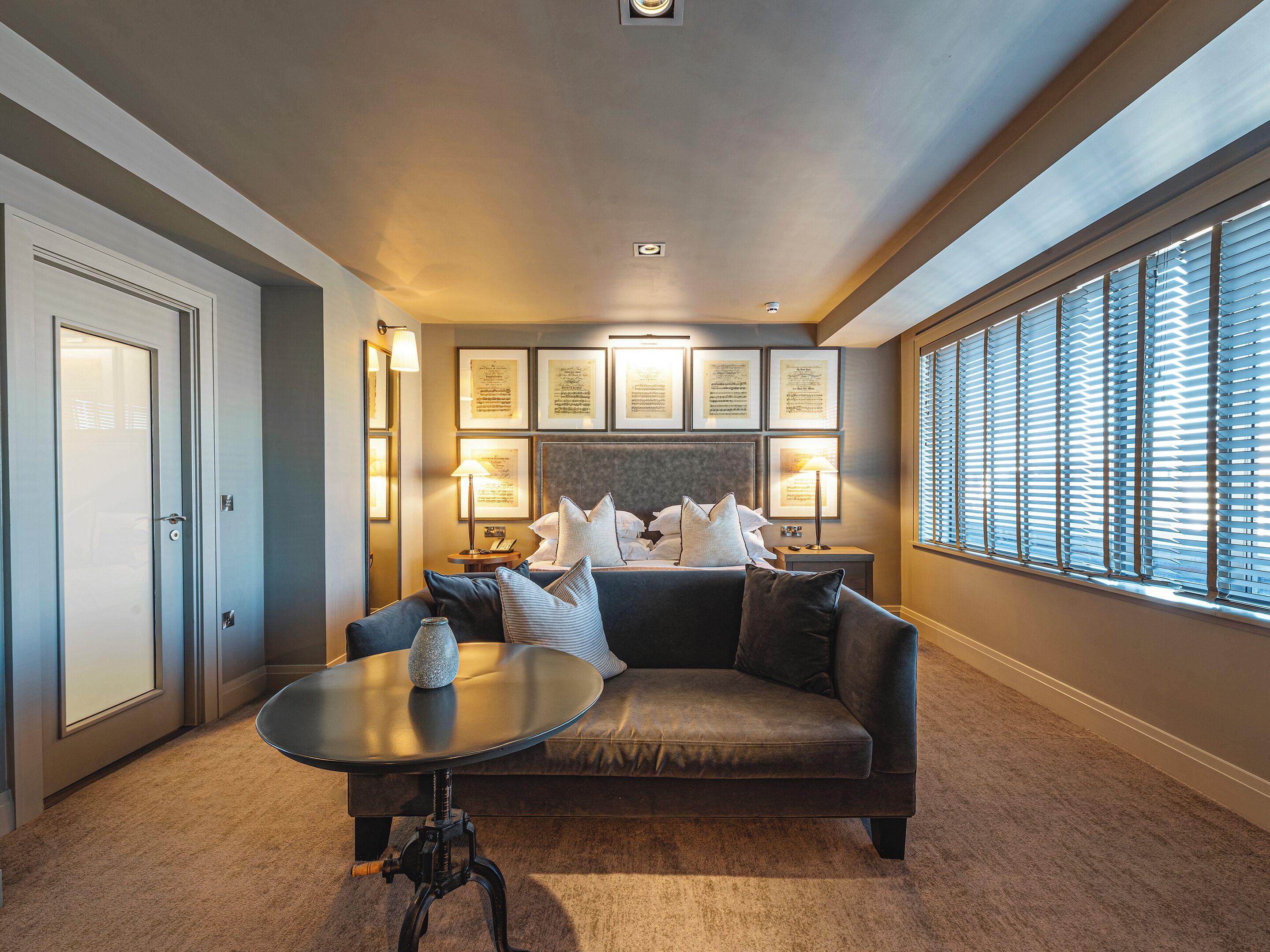 Habitación de hotel con sofá, una mesita, cama con almohadas, certificados enmarcados en la pared y ventana con persianas.