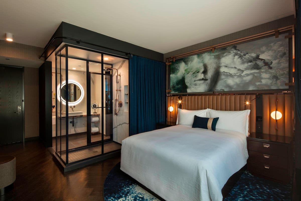 A modern hotel room with a large bed, a glass-enclosed shower area, a large painting on the wall, and a circular light fixture above the bed.