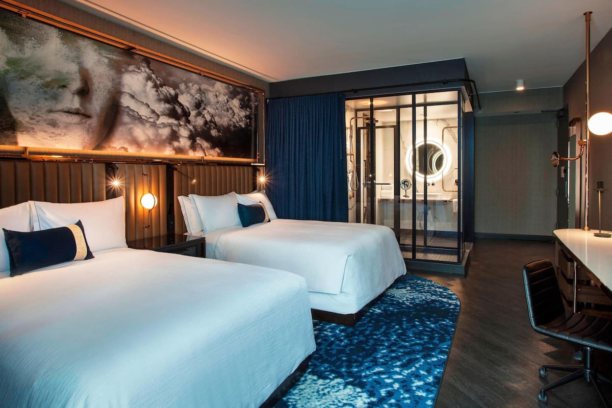 A hotel room with two beds, a large painting, a glass door leading to a bathroom, and a desk with a chair.
