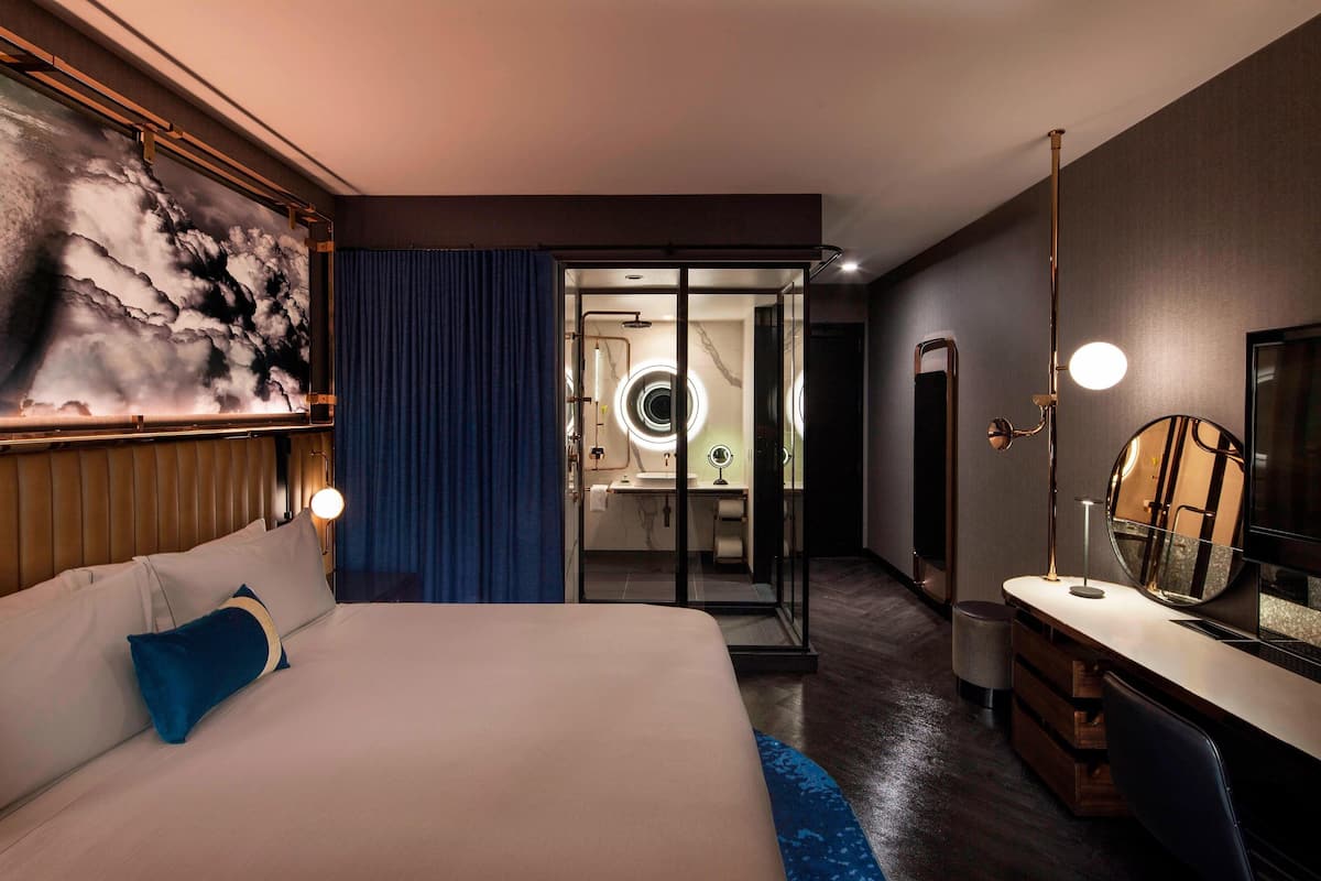 A hotel room with a bed, a desk with a mirror, a TV, and a bathroom with a glass door.