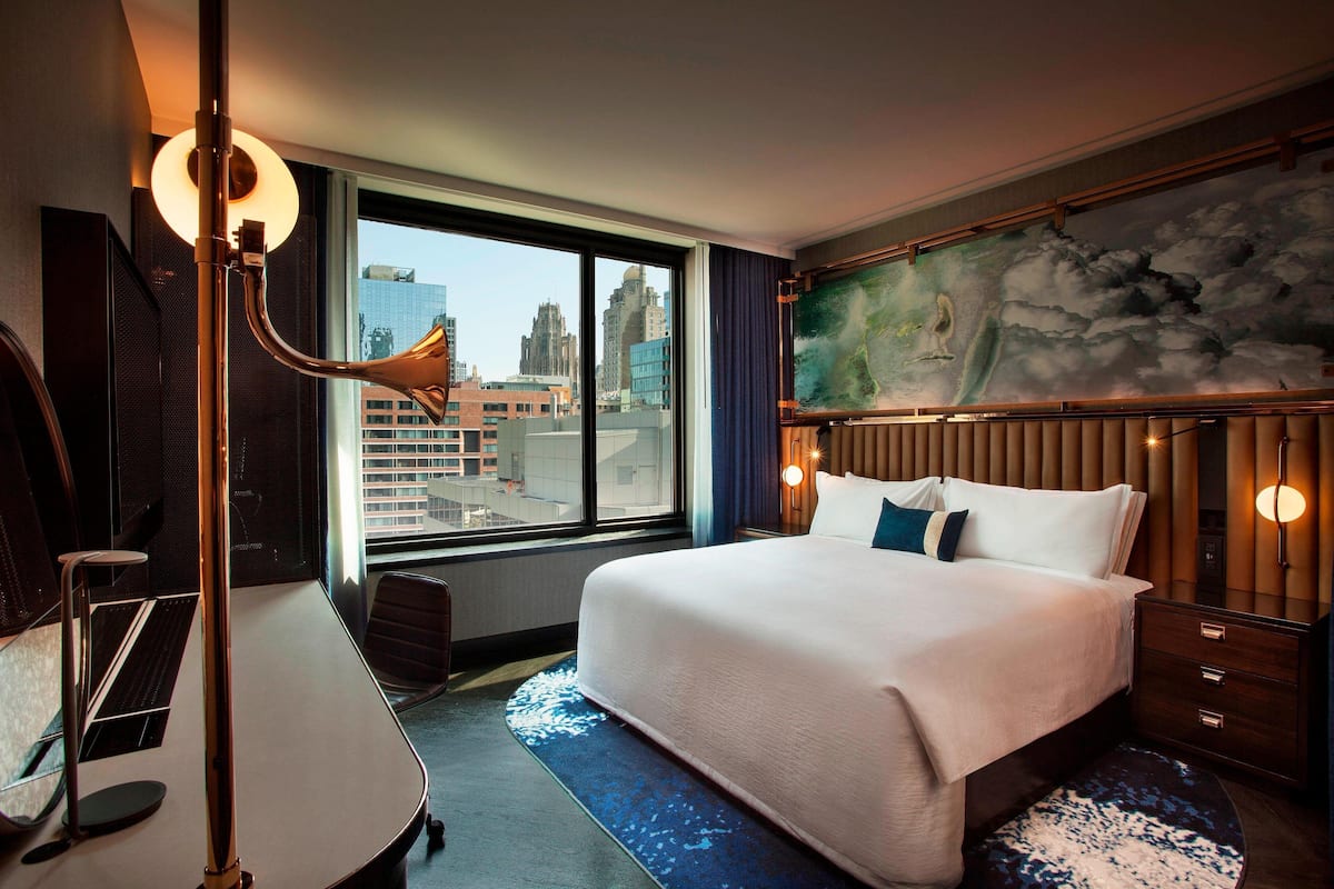 A hotel room with a large bed, a desk with a computer, a view of the city through the window, and a painting on the wall.