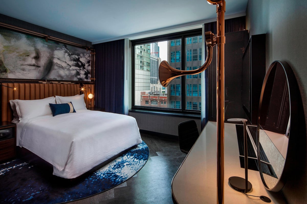 A modern hotel room with a large bed, a desk, a standing lamp, and a large window with a city view.