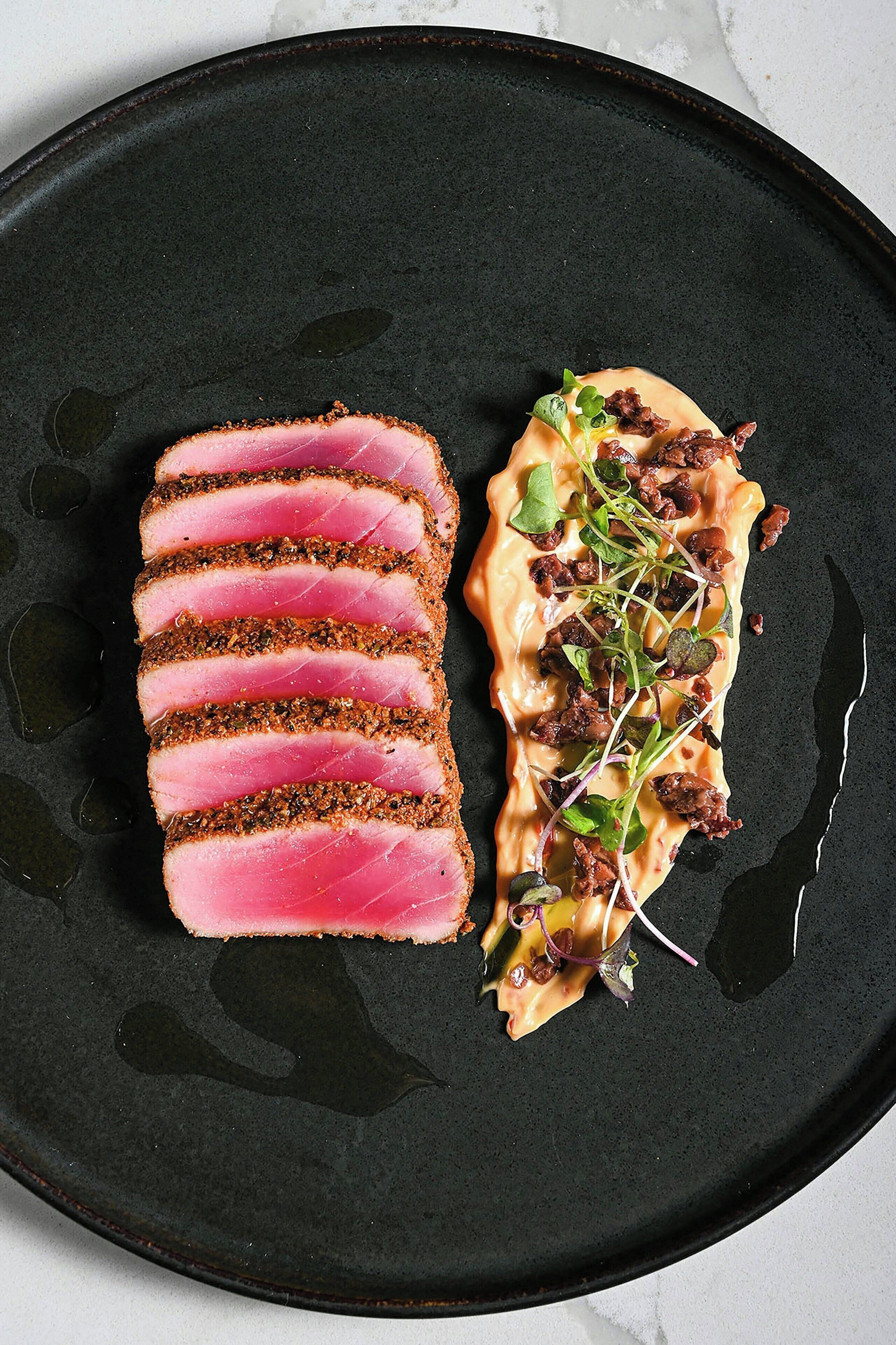 A black plate with sliced pink meat and a creamy sauce with greens and nuts.